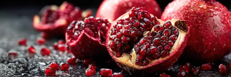 Close-up of a split pomegranate revealing juicy red seeds, with whole pomegranates and scattered seeds in a dark, glossy background.